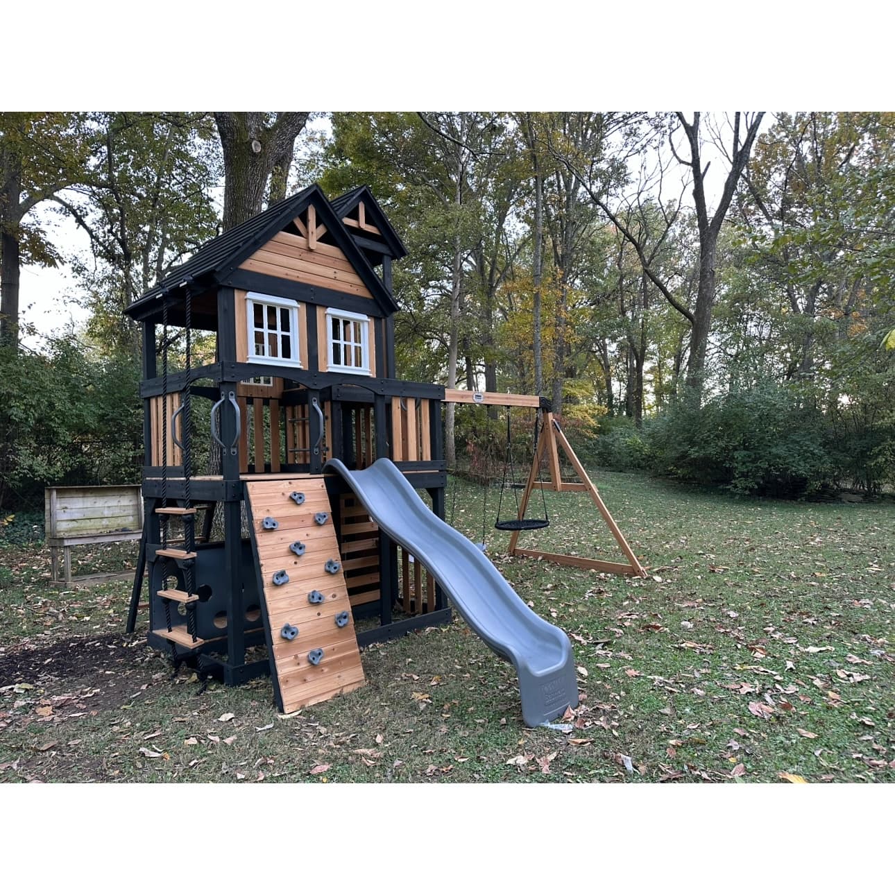 Cedar and black playset fort with a slide, rock wall, climbing rope and swing beam, built in a wooded backyard